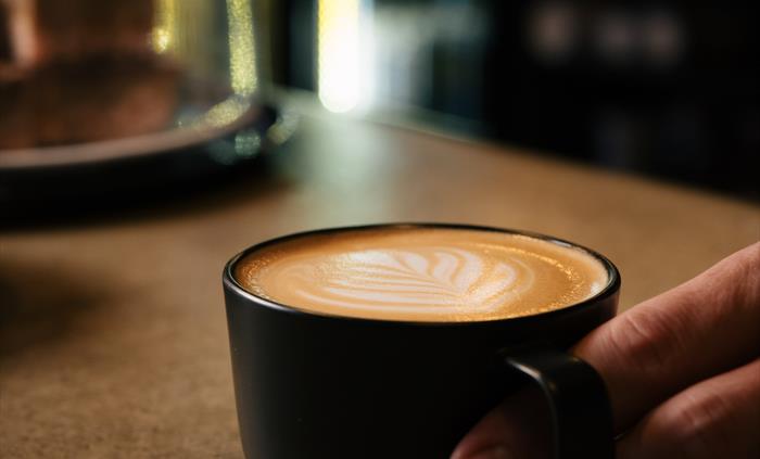A dark, low coffee cup sits on a concrete bar with a leaf motif decorating the top of the coffee. A hand to the right of the image has a finger throug